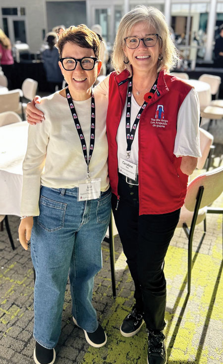 Two adult female arm amputees stand in a dining room with their arms around each other.