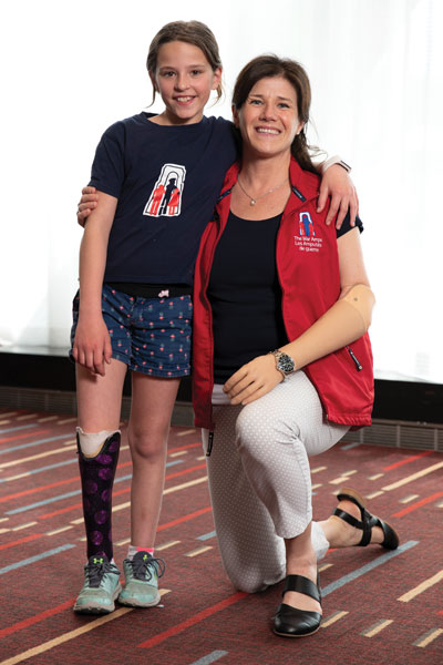 Une enfant amputée d’une jambe se tient debout avec son bras autour d’une femme amputée d’un bras qui est agenouillée pour être à sa hauteur.
