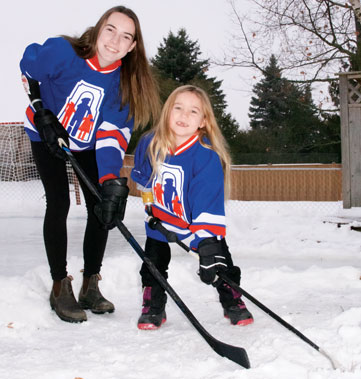 Two young female arm amputees pose together on an outdoor ice rink while holding hockey sticks.
