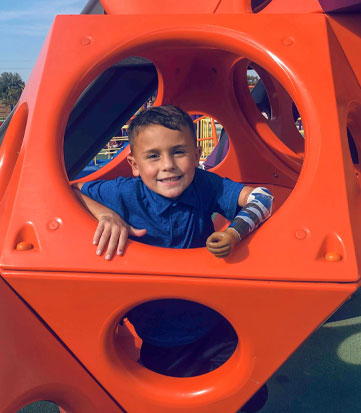 A young male arm amputee wearing his artificial arm peeks out from inside a play structure at a playground.