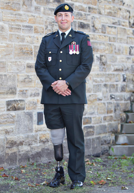An adult male veteran with a leg amputation stands in uniform beside a stone wall.
