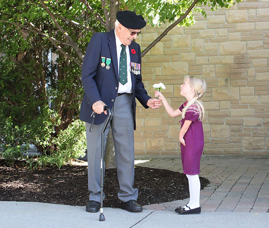 A young female upper limb amputee hands a flower to a Second World War veteran with a leg amputation.