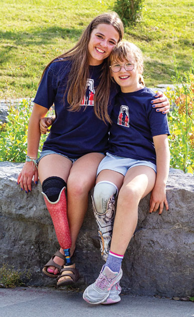 A young adult female leg amputee sits on a large rock in a park with her arm around a young female leg amputee.