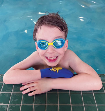 A young male partial hand amputee wears swimming goggles and rests his arms on the edge of the swimming pool.
