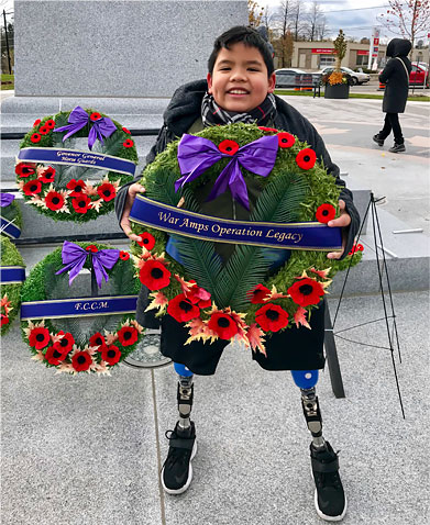 A young male double leg amputee holding a Remembrance Day wreath standing in front of a cenotaph.