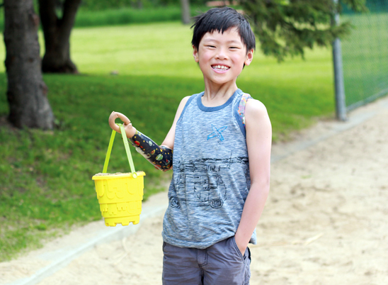 Un jeune garçon amputé d’un bras sur un terrain de jeu tient un seau à sable jaune avec son membre artificiel.
