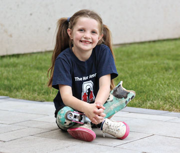 A young female double leg amputee sits cross-legged on the ground outside.