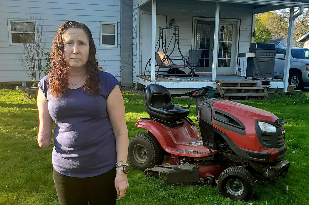 A woman who is missing her right arm above the elbow stands outside in front of a riding lawn mower.
