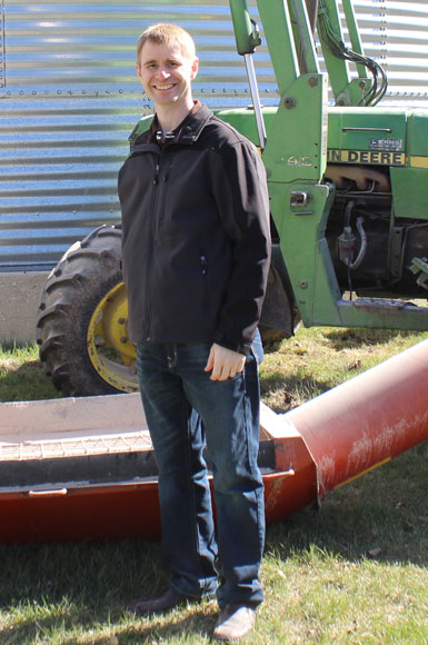Merrill, a right arm amputee, stands in front of a grain auger on a farm.