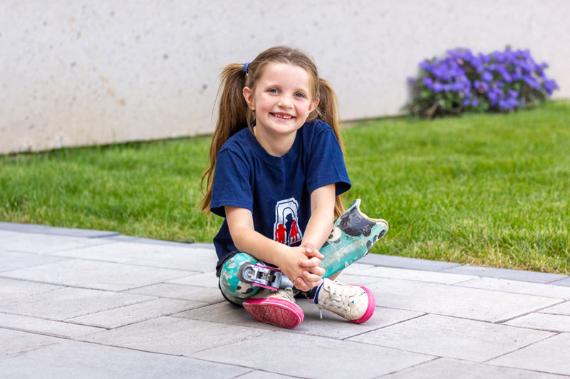 A young girl wearing artificial legs sitting on the pavement with her legs crossed.