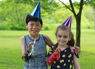 Deux jeunes amputés à un bras, debout dans un parc avec les bras enlacés l’un autour de l’autre, portent des chapeaux de fête et tiennent des cornes de fête.