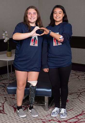 Two young girls, one an arm amputee and the other a leg amputee, join their hands together to create the shape of a heart.