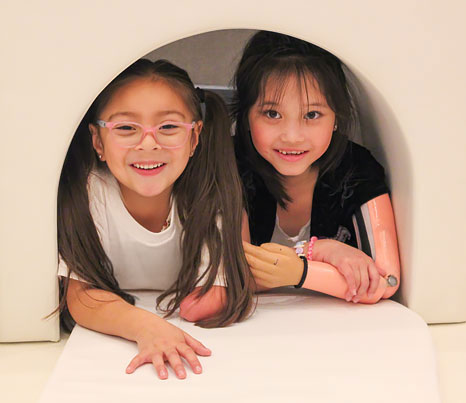 Two young girls, both arm amputees, play inside a white foam structure.